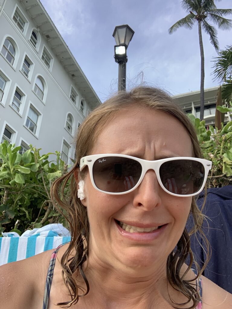 peach skinned woman wearing white sunglasses grimacing, with visible tissue in ear